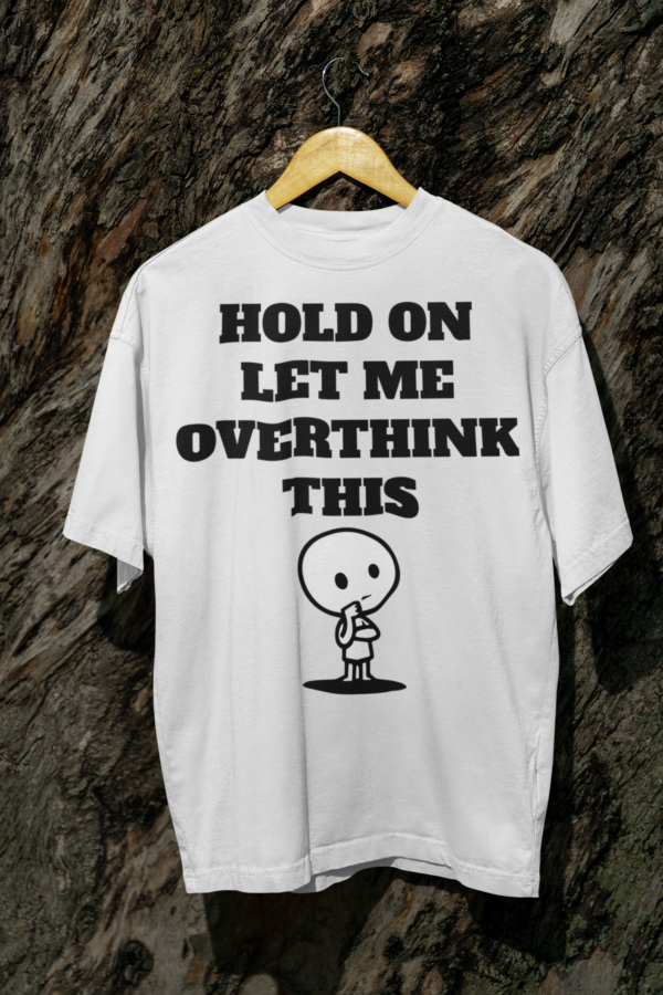 A large white T-shirt with the text "Hold On Let Me Overthink This" hanging on a tree log, featuring a cute cartoon character in a thoughtful pose.